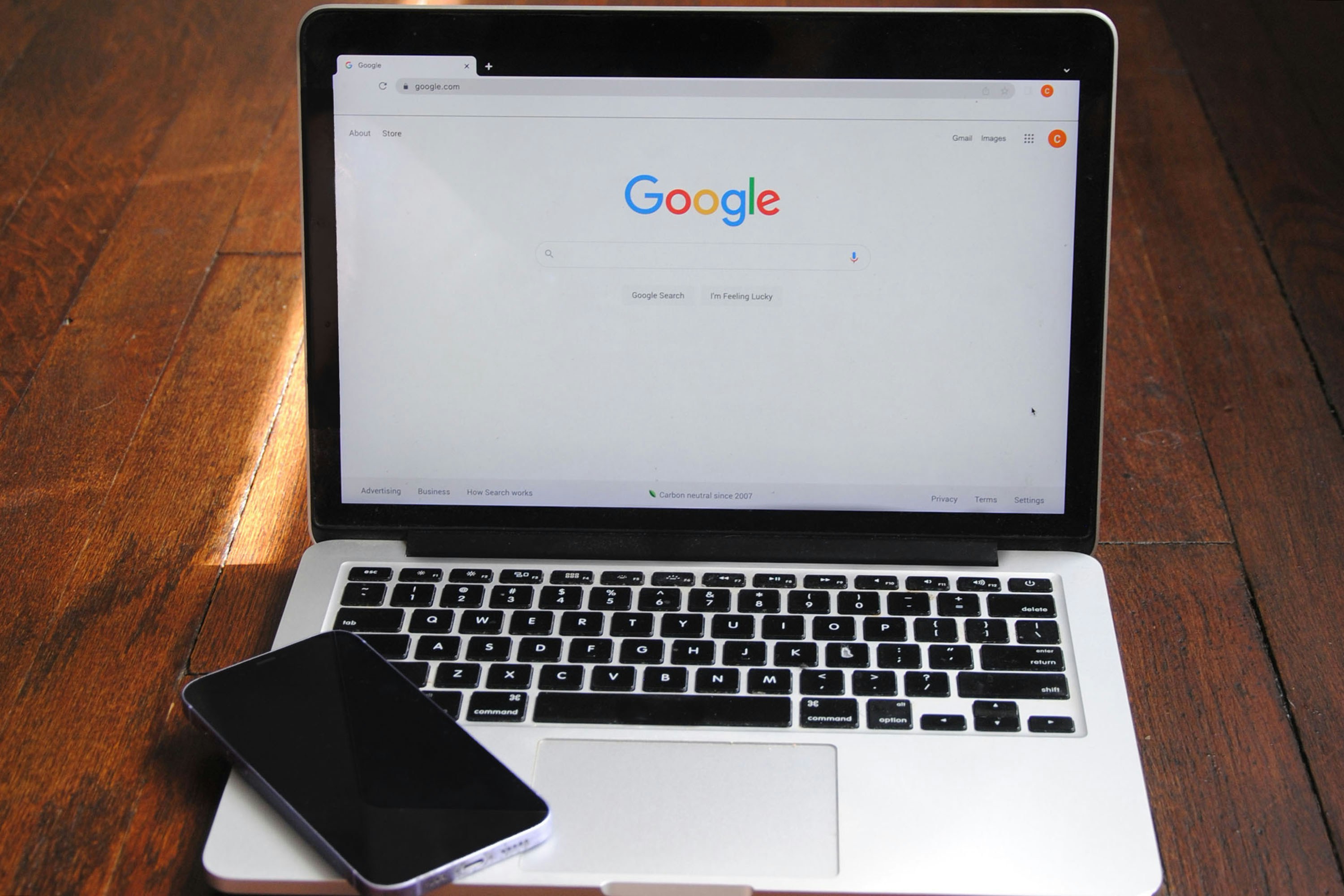 MacBook Pro ouvert sur la page d'accueil Google, posé sur un bureau en bois avec un smartphone. Illustration article blog optimisation fiche établissement Google